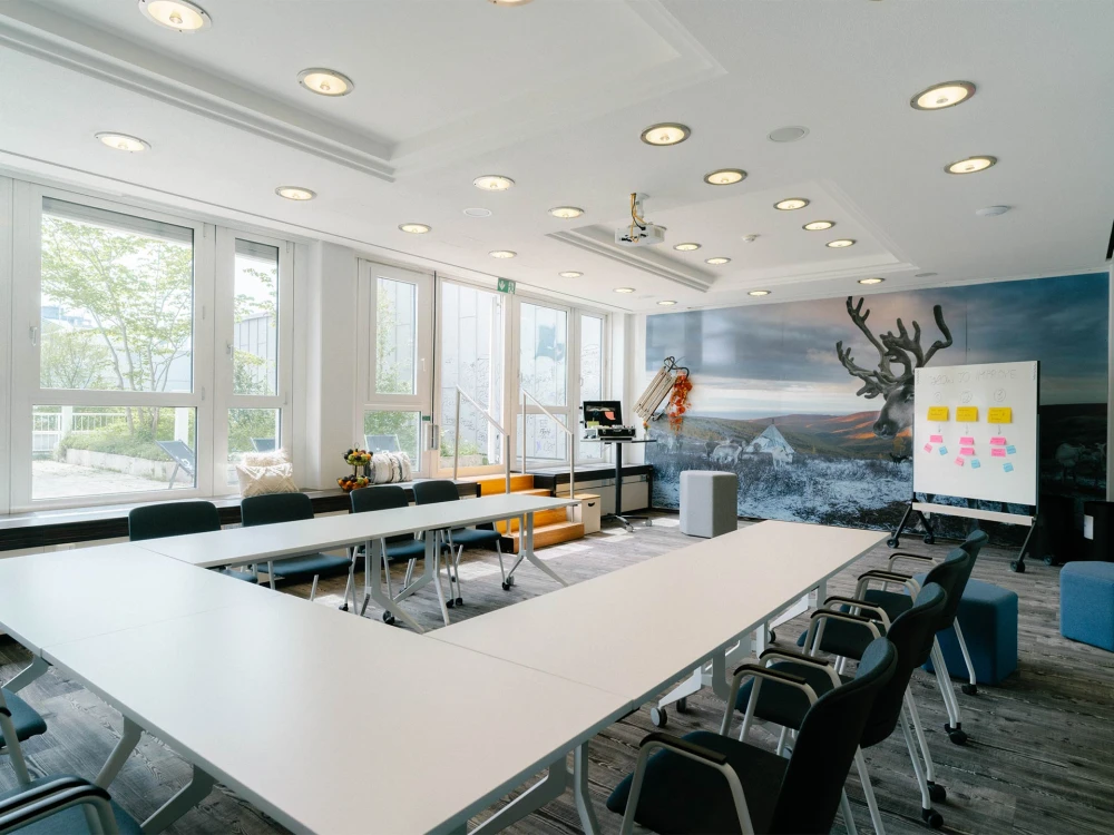 Heller, moderner Meetingraum mit großen Fenstern, U-förmiger Tischanordnung, Whiteboard mit Notizen und einer Wandgestaltung mit Naturmotiv.