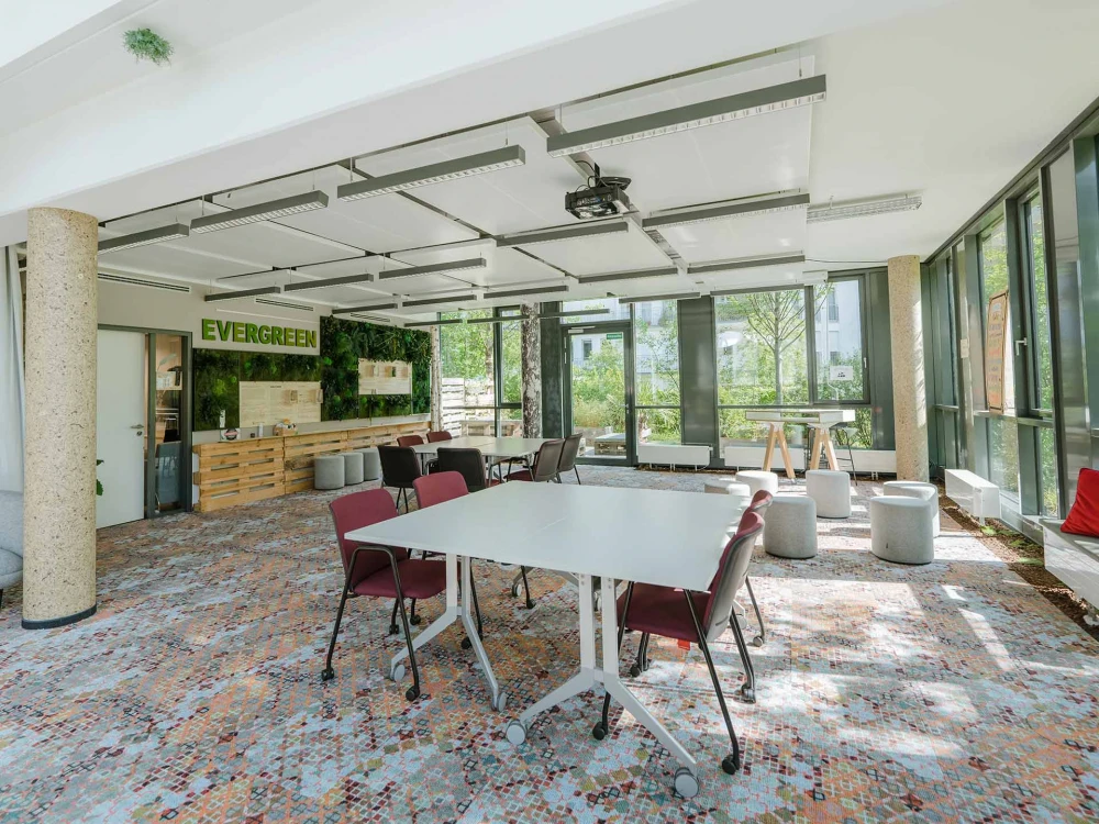 Heller, moderner Workshopraum mit großen Fenstern, flexibler Möblierung, grüner Wandgestaltung und gemütlicher Atmosphäre, ideal für kreative Meetings.