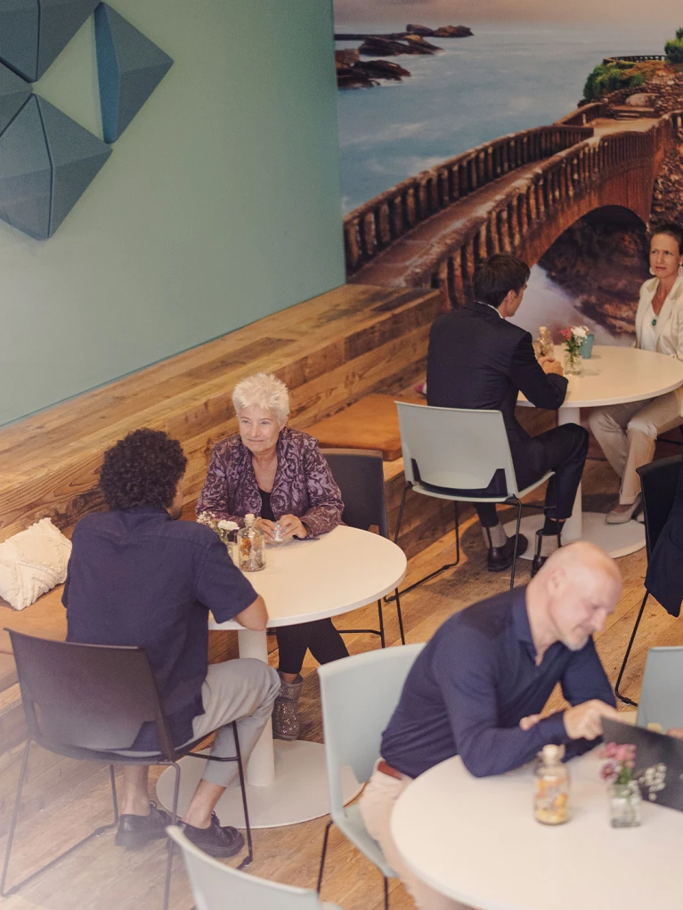 Moderne Café- oder Coworking-Umgebung mit Menschen, die an runden Tischen Gespräche führen. Im Hintergrund eine große Wand mit einer Landschaftsfotografie.