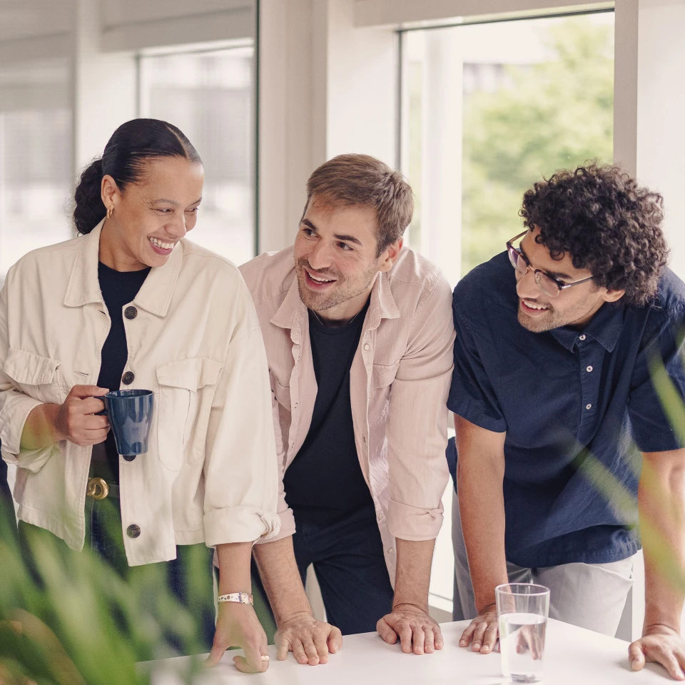 Drei Kollegen in einem modernen Büro stehen an einem Tisch, lachen und unterhalten sich entspannt. Eine Frau hält eine Kaffeetasse, im Vordergrund grüne Pflanzen.