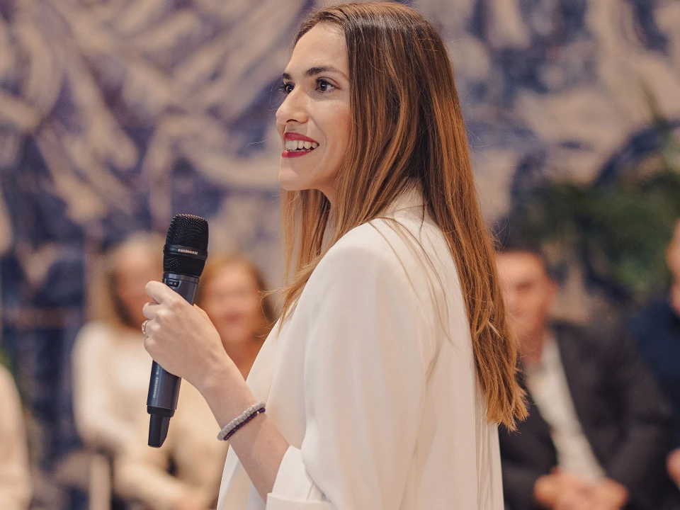 Junge Frau in einem weißen Blazer hält ein Mikrofon und spricht vor einem Publikum in einem modernen Veranstaltungsraum mit kunstvoller Wandgestaltung.