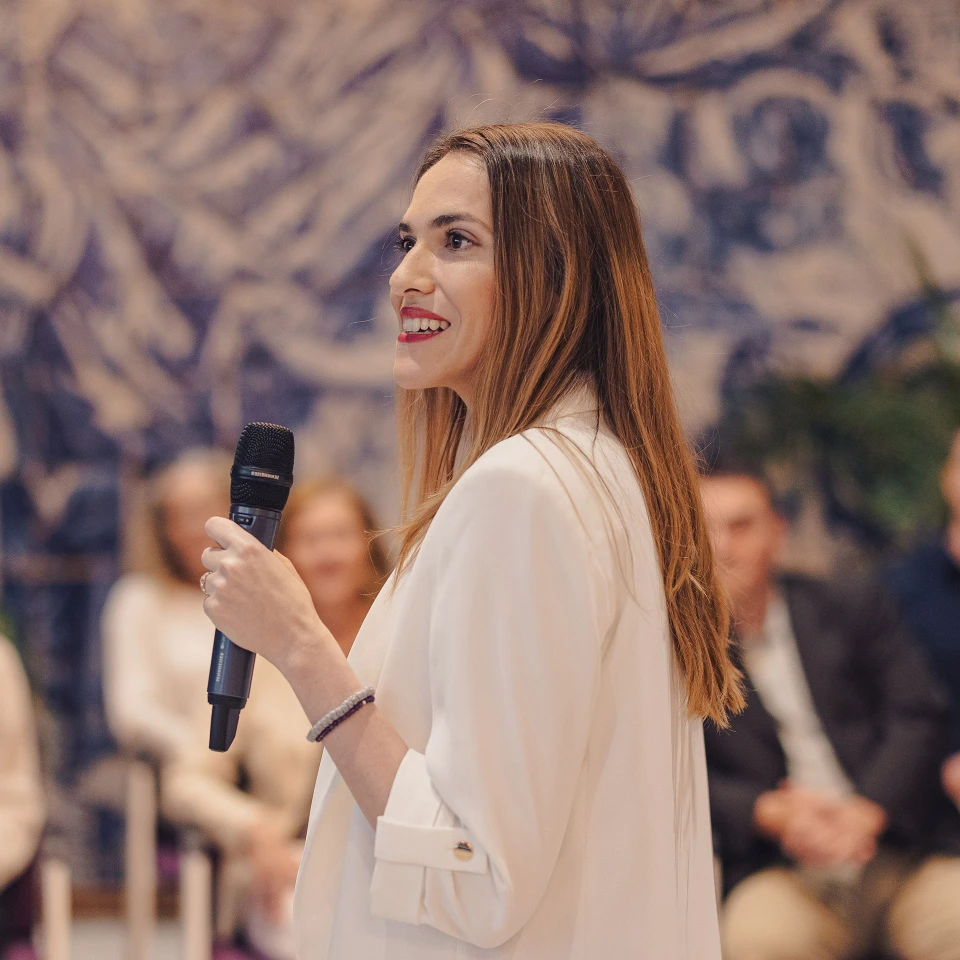 Junge Frau in einem weißen Blazer hält ein Mikrofon und spricht vor einem Publikum in einem modernen Veranstaltungsraum mit kunstvoller Wandgestaltung.
