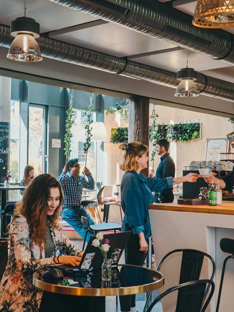 Modernes Café mit urbanem Design, in dem Menschen Kaffee genießen, arbeiten und sich unterhalten. Ein Barista serviert einer Kundin einen Kaffee an der Theke.
