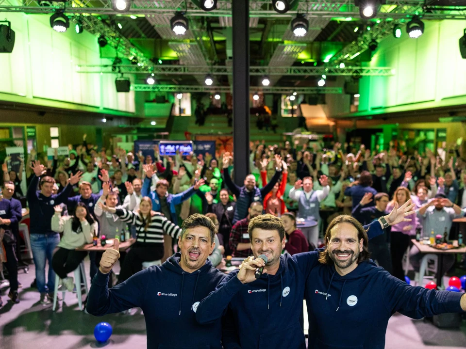 Drei Moderatoren auf einer Bühne mit Mikrofonen vor einer großen, begeisterten Menschenmenge bei einem Event in einer modern beleuchteten Veranstaltungshalle.