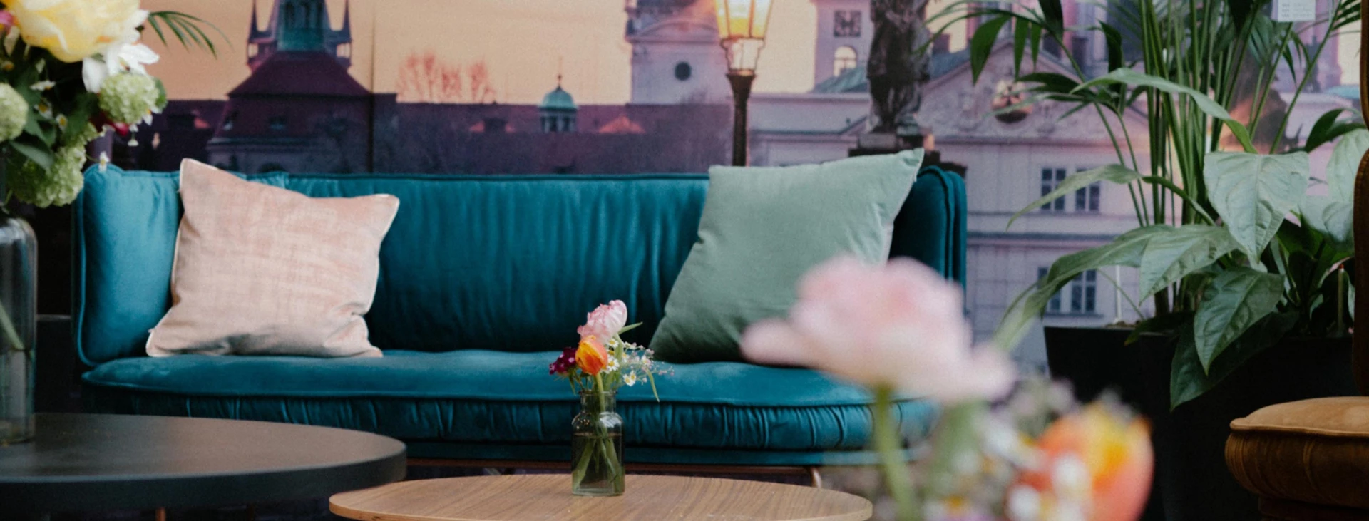 Gemütliche Lounge-Ecke mit einer türkisfarbenen Samtcouch, Zierkissen, Blumen auf einem Holztisch und einer Wandgestaltung mit Stadtmotiv und Pflanzen.