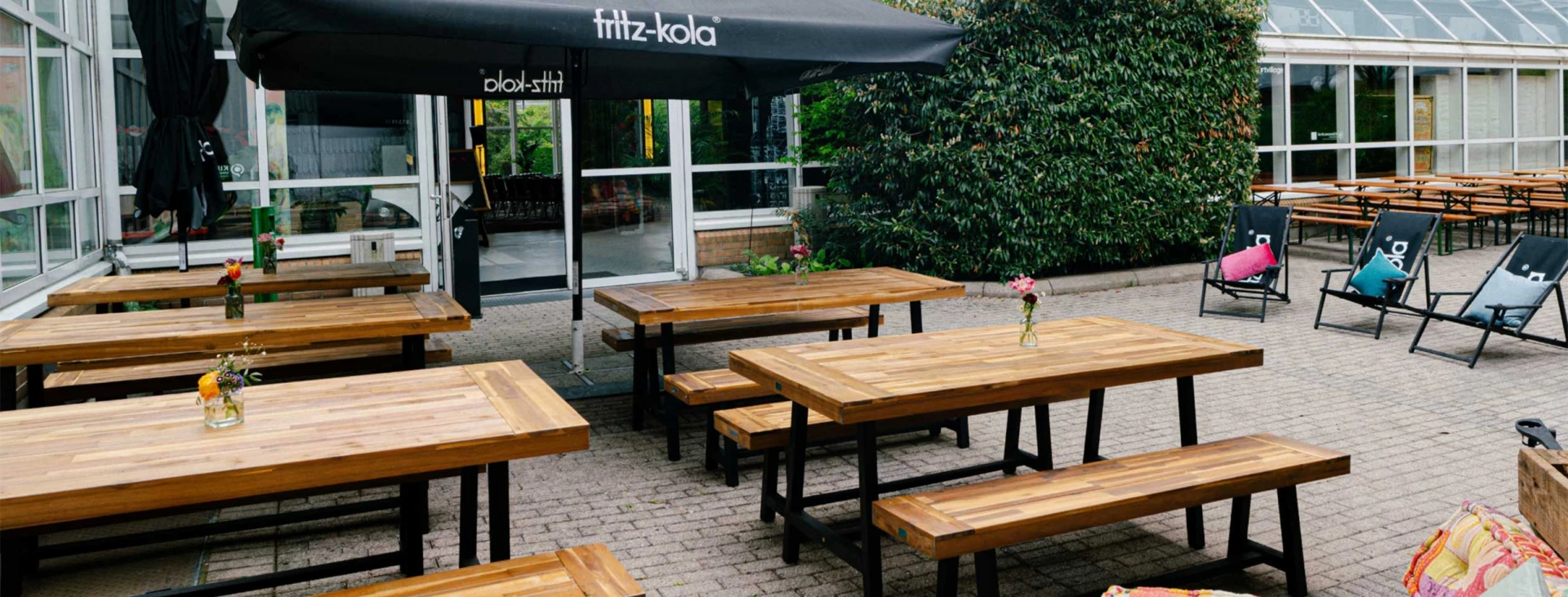Gemütlicher Außenbereich mit Holzbänken und Tischen, dekoriert mit kleinen Blumenvasen. Sonnenliegen mit bunten Kissen und ein Fritz-Kola-Sonnenschirm sorgen für eine entspannte Atmosphäre.