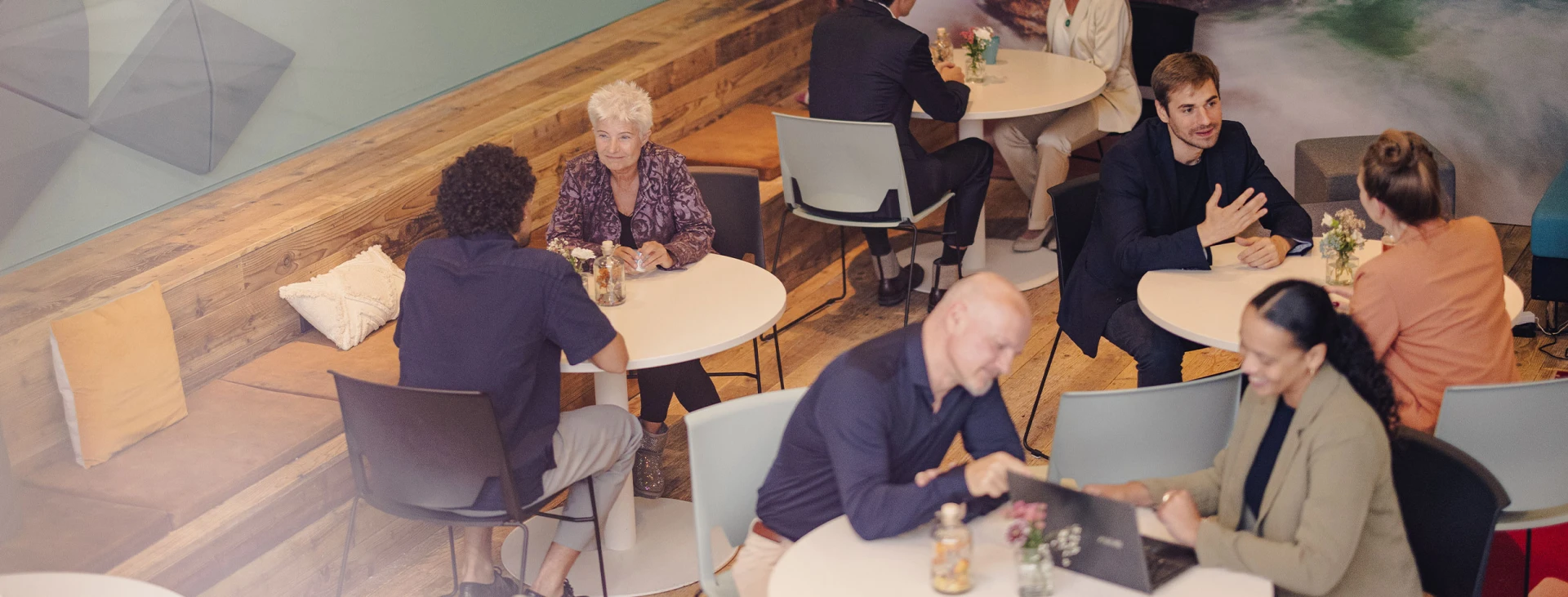 Moderne Café- oder Coworking-Umgebung mit Menschen, die an runden Tischen Gespräche führen. Im Hintergrund eine große Wand mit einer Landschaftsfotografie.