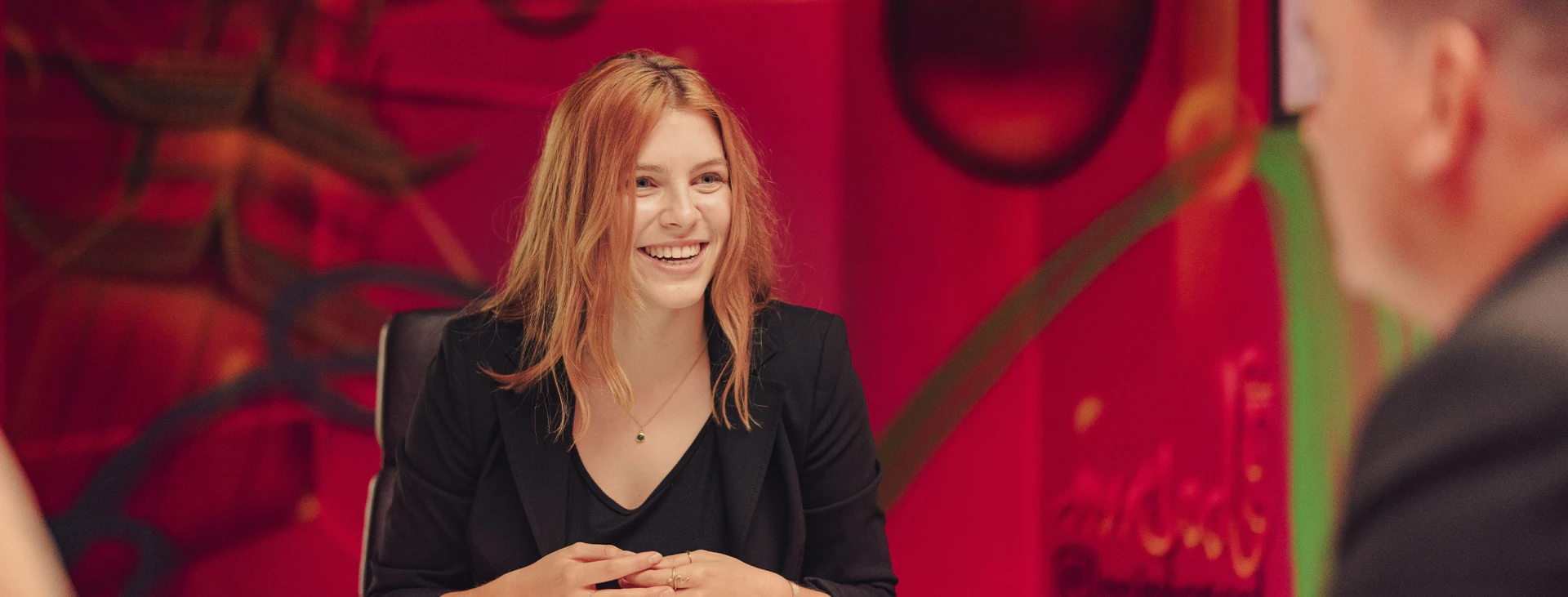 Junge Frau mit roten Haaren lacht während eines Gesprächs in einem modernen Meetingraum mit kreativ gestalteter, roter Wand im Hintergrund.