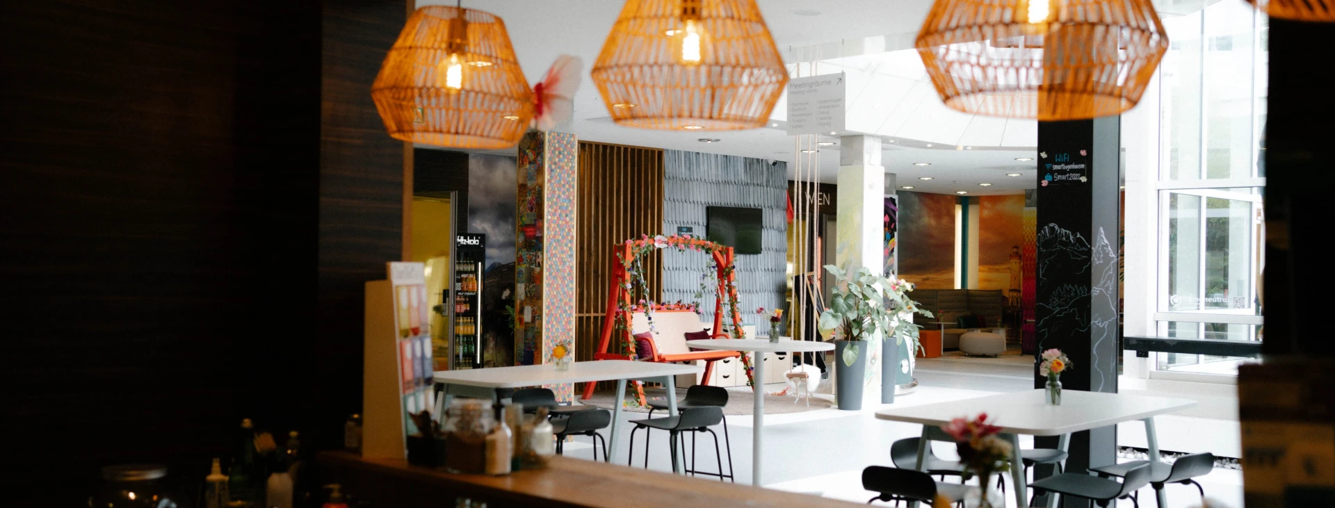 Blick aus einer Café-Theke auf einen modernen, stilvoll dekorierten Coworking- oder Lounge-Bereich mit hängenden Lampen, Pflanzen und Sitzgelegenheiten.