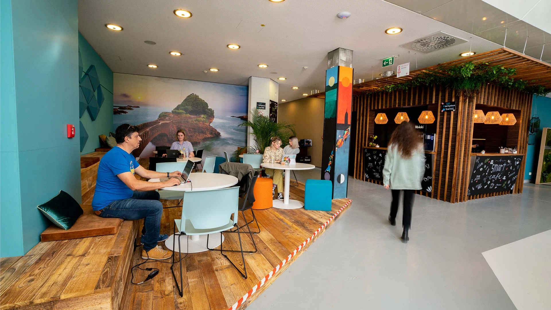 Modernes Café mit kreativer Einrichtung, Holz- und Pflanzen-Elementen. Menschen arbeiten an Laptops und unterhalten sich an runden Tischen. Barista-Bereich im Hintergrund.