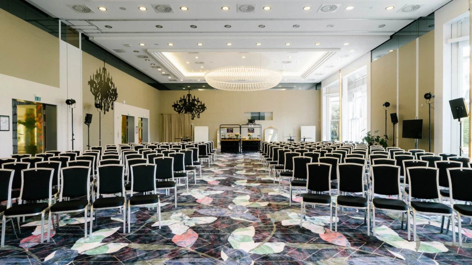 Eleganter, moderner Konferenzraum mit Reihenbestuhlung für ein Event oder eine Tagung. Die schwarzen Stühle mit weißen Rahmen sind symmetrisch ausgerichtet und führen zu einer Bühne im Hintergrund, die mit einem Tisch, Whiteboards und Dekorationen ausgestattet ist. Die hohen Decken mit einem großen Kronleuchter sorgen für eine edle Atmosphäre. Der Boden ist mit einem farbenfrohen Teppichmuster gestaltet, während große Fenster auf der rechten Seite viel Tageslicht hereinlassen. An den Seitenwänden befinden sich dekorative Spiegel und Lichtinstallationen für eine stimmungsvolle Beleuchtung