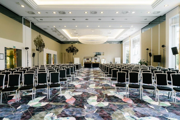 Eleganter, moderner Konferenzraum mit Reihenbestuhlung für ein Event oder eine Tagung. Die schwarzen Stühle mit weißen Rahmen sind symmetrisch ausgerichtet und führen zu einer Bühne im Hintergrund, die mit einem Tisch, Whiteboards und Dekorationen ausgestattet ist. Die hohen Decken mit einem großen Kronleuchter sorgen für eine edle Atmosphäre. Der Boden ist mit einem farbenfrohen Teppichmuster gestaltet, während große Fenster auf der rechten Seite viel Tageslicht hereinlassen. An den Seitenwänden befinden sich dekorative Spiegel und Lichtinstallationen für eine stimmungsvolle Beleuchtung