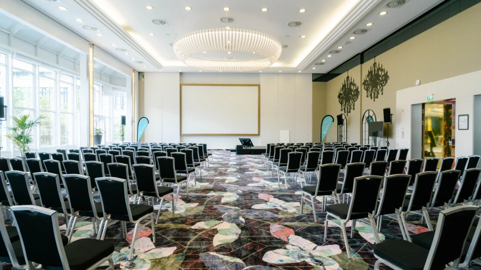 Heller und stilvoller Konferenzsaal mit moderner Bestuhlung, großem Projektionsbildschirm und eleganter Beleuchtung. Perfekt für Tagungen, Präsentationen und Business-Events