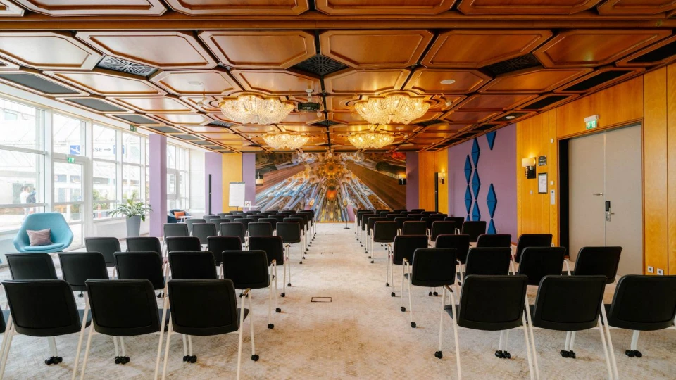 Modern eingerichteter Konferenzraum mit einer warmen Holzdecke, luxuriösen Kronleuchtern und einer beeindruckenden Wandgestaltung im futuristischen Stil. Die Reihen mobiler schwarzer Stühle sind auf eine Präsentationsfläche ausgerichtet, während große Fenster für viel Tageslicht sorgen. Der Raum verbindet klassisches Design mit innovativen Akzenten und eignet sich ideal für Tagungen, Workshops und Events