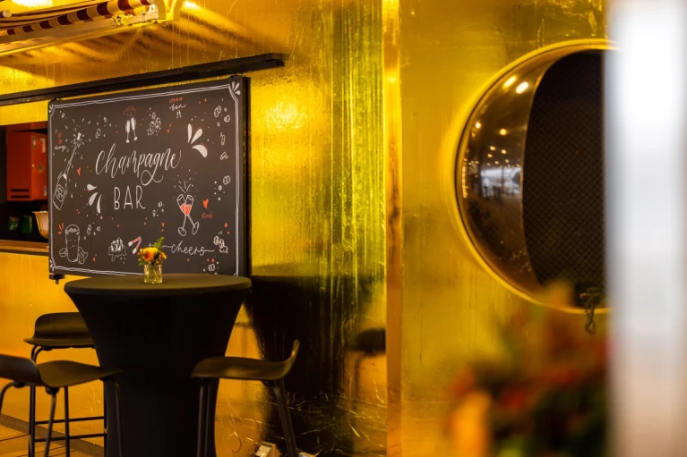 Champagner-Bar mit goldener Wandverkleidung, schwarzem Stehtisch und Hockern. Eine handbeschriftete Tafel mit der Aufschrift 'Champagne Bar' und dekorativen Illustrationen hängt an der Wand. Rechts spiegelt sich der Raum in einer runden, metallischen Öffnung.