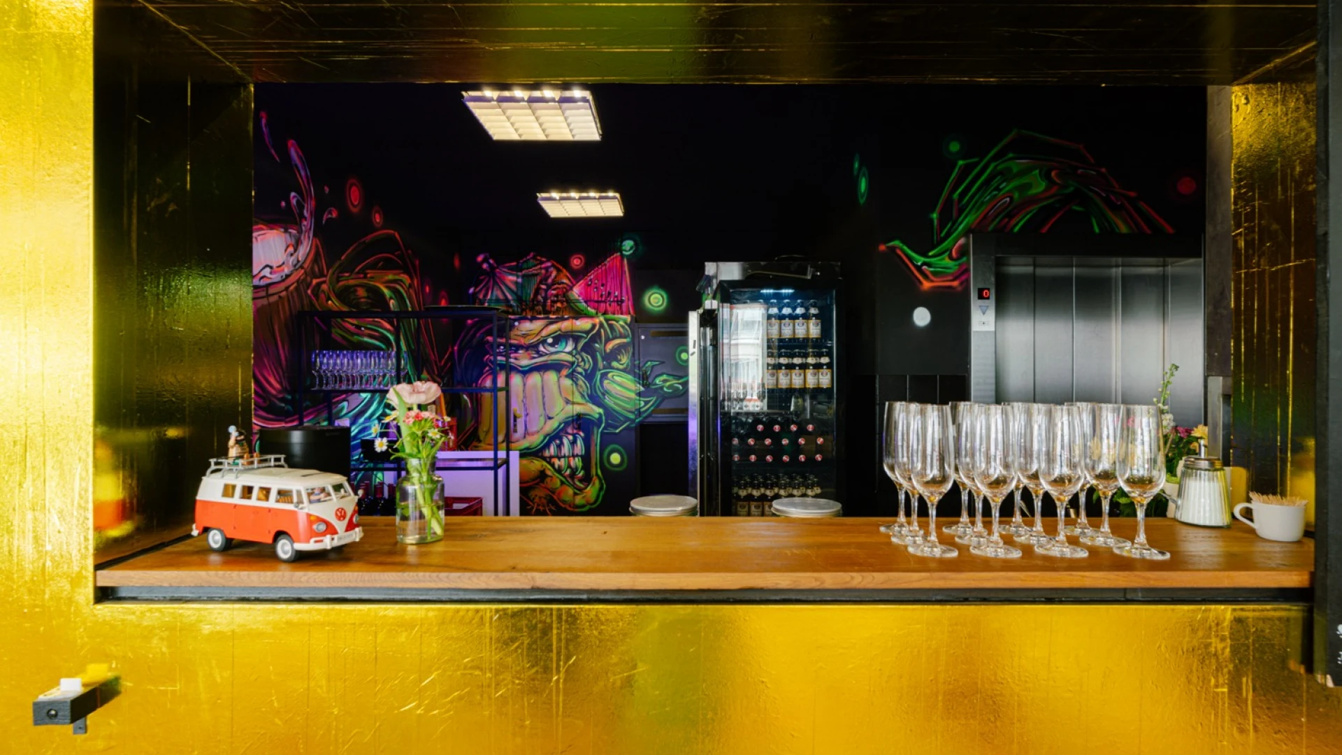 Champagner-Bar mit goldener Wandverkleidung und einer Durchreiche. Auf der Theke stehen aufgereihte Sektgläser, eine kleine Blumenvase und ein Modellbus. Im Hintergrund eine bunt beleuchtete Bar mit neonfarbenem Graffiti, einem Getränkekühlschrank und Regalen mit Gläsern.