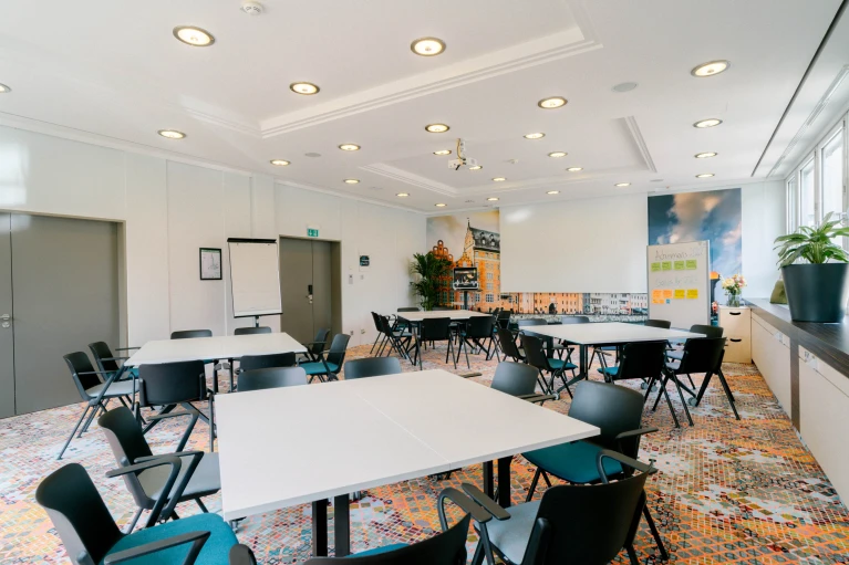 Heller und moderner Konferenzraum mit mehreren weißen Tischen und schwarzen Stühlen, angeordnet für Gruppenarbeit. An der Wand ein großes Stadtpanorama mit historischen Gebäuden. Eine Flipchart und ein Whiteboard mit Notizen stehen bereit. Große Fenster auf der rechten Seite lassen viel Tageslicht herein, während grüne Pflanzen den Raum ergänzen