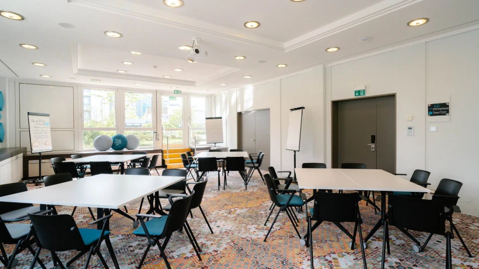 Moderner Meetingraum mit weißen Tischen, schwarzen Stühlen, Flipcharts und großen Fenstern mit Blick ins Freie