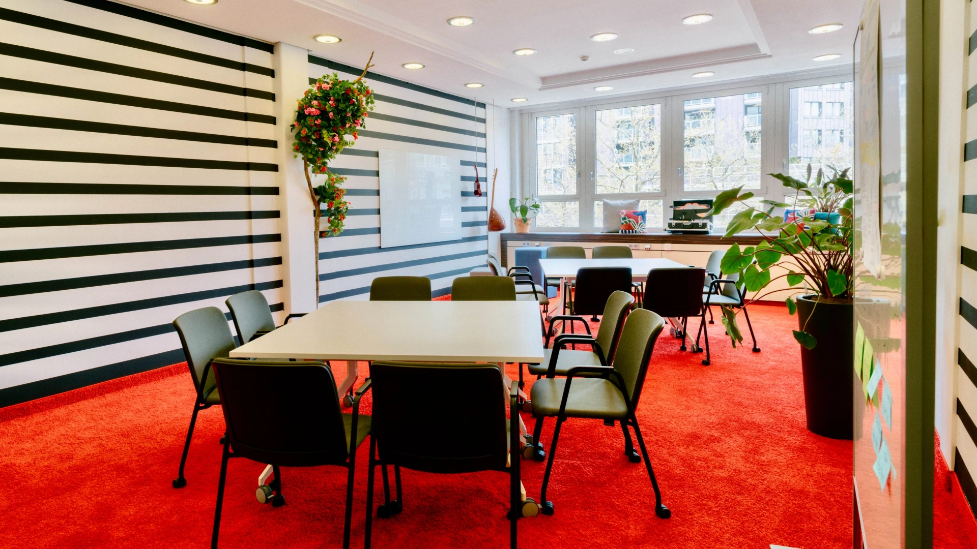 Meetingraum mit rotem Teppich, gestreiften Wänden, weißen Tischen, schwarzen Stühlen und großen Fenstern mit Tageslicht.