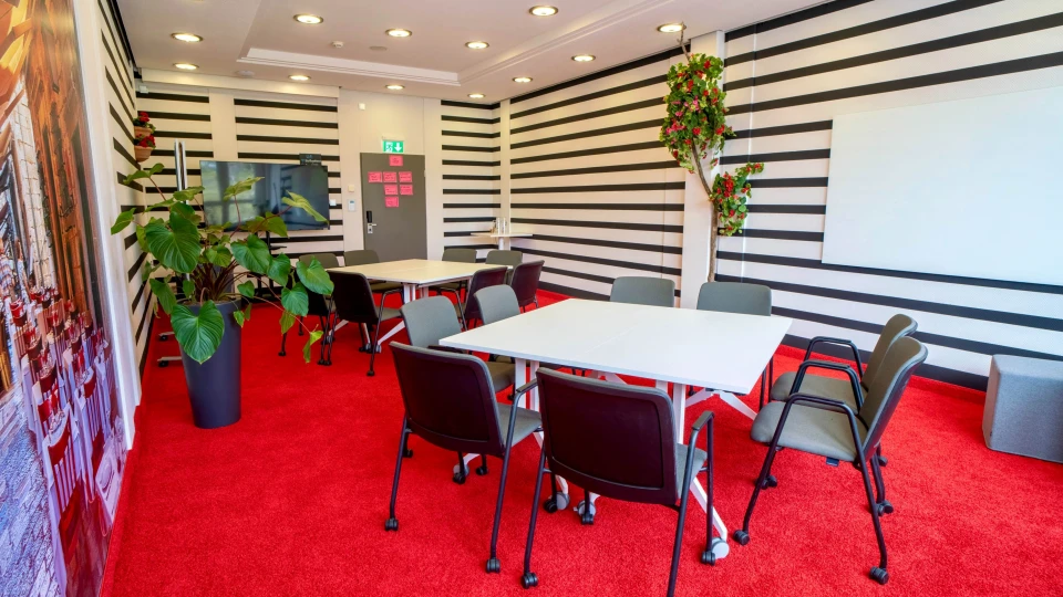 Moderner Konferenzraum mit schwarz-weiß gestreiften Wänden, roten Teppichen, weißen Tischen, Pflanzen und Wandtafel.