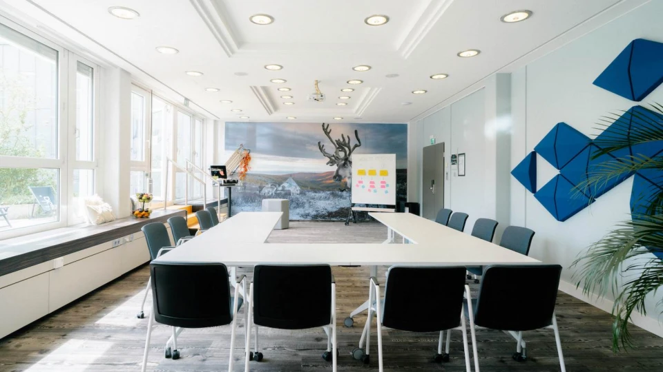 Moderner Konferenzraum mit U-förmiger Tischanordnung, großen Fenstern, Wandkunst mit Rentiermotiv und blauen Akustikpaneelen.