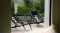 Blick durch ein Fenster auf eine Terrasse mit zwei schwarzen Liegestühlen, umgeben von grüner Bepflanzung.
