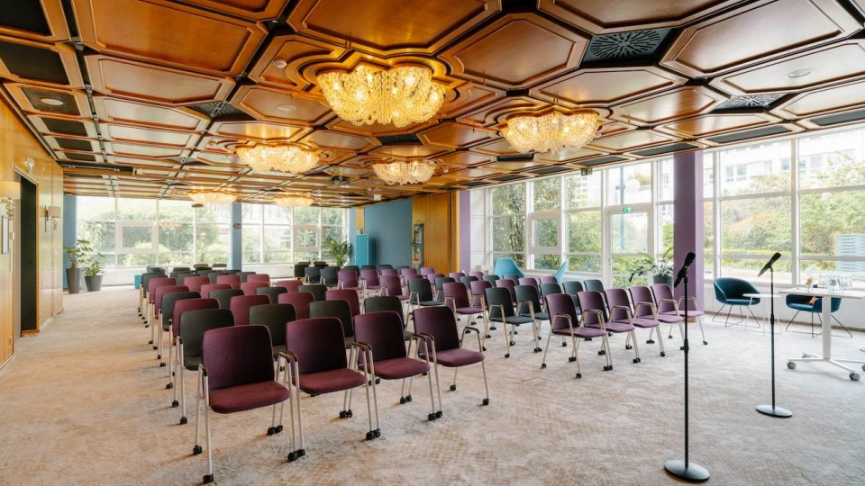 Konferenzraum mit lila und schwarzen Stühlen, Mikrofonen, großen Fenstern und Kronleuchtern an der Holzdecke