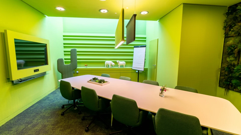 Konferenzraum mit grünem Design, runder Tisch mit Stühlen, Akustikpaneelen, Flipchart und Dekorationen
