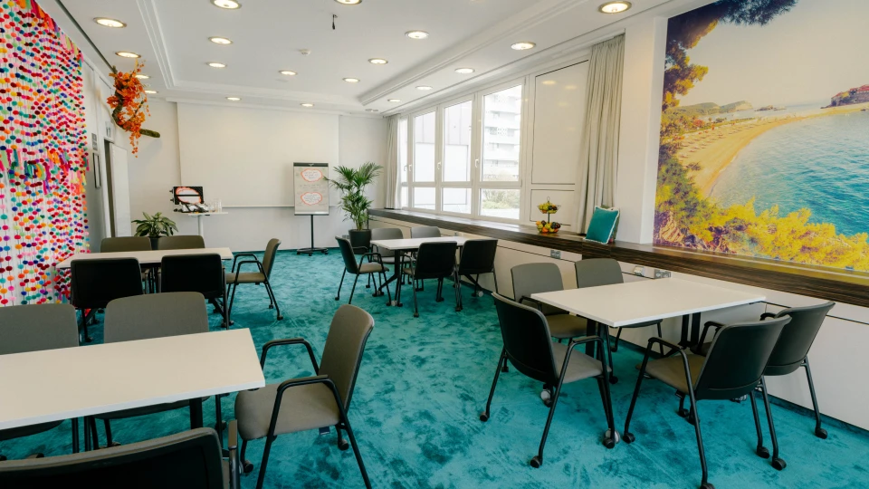Heller Meetingraum mit türkisfarbenem Teppich, weißen Tischen und grauen Stühlen. Die Wand ist mit einem großen Strandmotiv sowie bunten Girlanden dekoriert, die eine kreative Atmosphäre schaffen