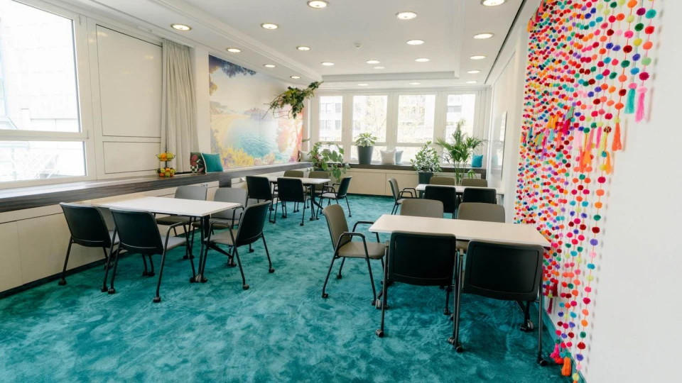 Heller Tagungsraum mit blauem Teppich, bunten Pompon-Girlanden und Pflanzen für eine kreative Arbeitsatmosphäre