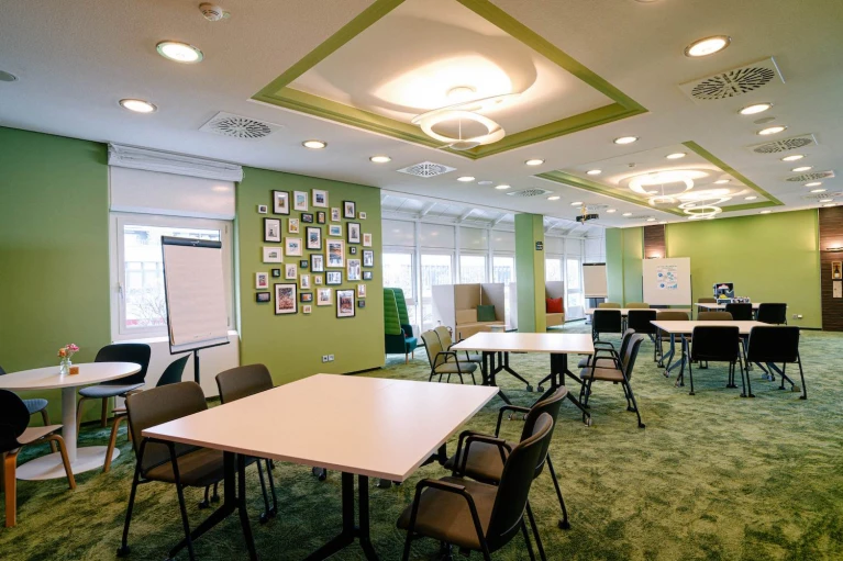 Meetingraum mit eckigen Tischen, türkisen Stühlen, grüner Wand und mediterranen Deko-Elementen.