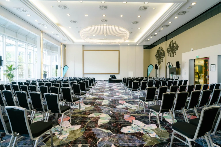 Symmetrischer Blick auf den Konferenzraum Wien mit zentraler Projektionsfläche, LED-Kronleuchter und großer Fensterfront.